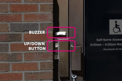 Door buzzer and up/down button on a brick wall next to a door with accessibility information.