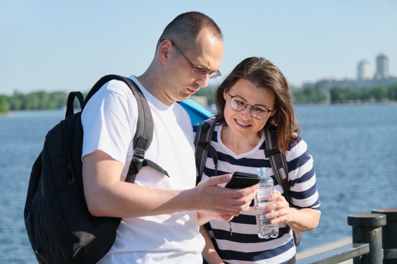 A man and woman with backpacks look at a smartphone together by a body of water.