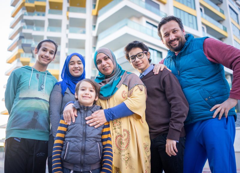 A smiling family of six, including parents and four children, poses for a photo in front of a modern apartment building.
